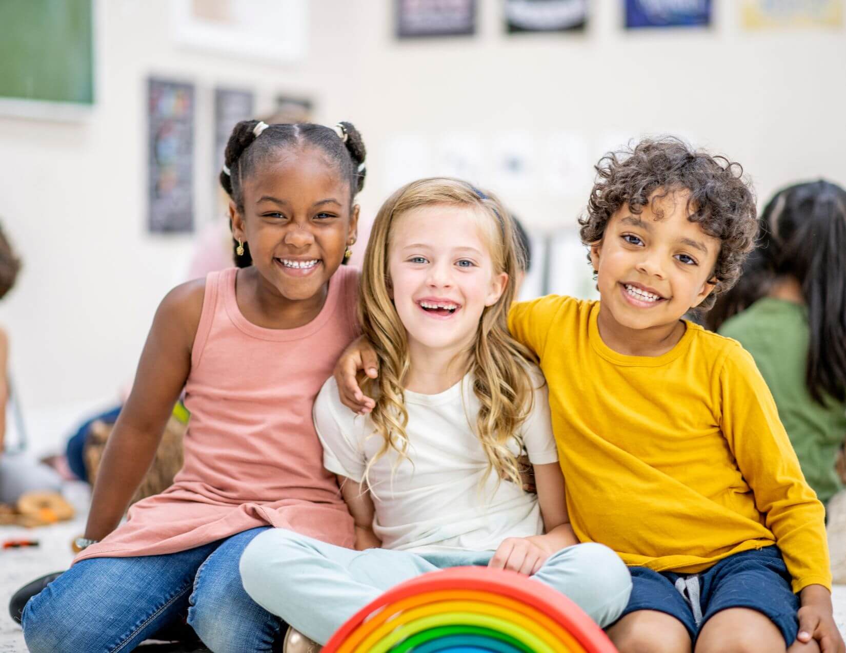 Body Space Awareness Three young children of various skin tones smile while sitting close together on the floor.