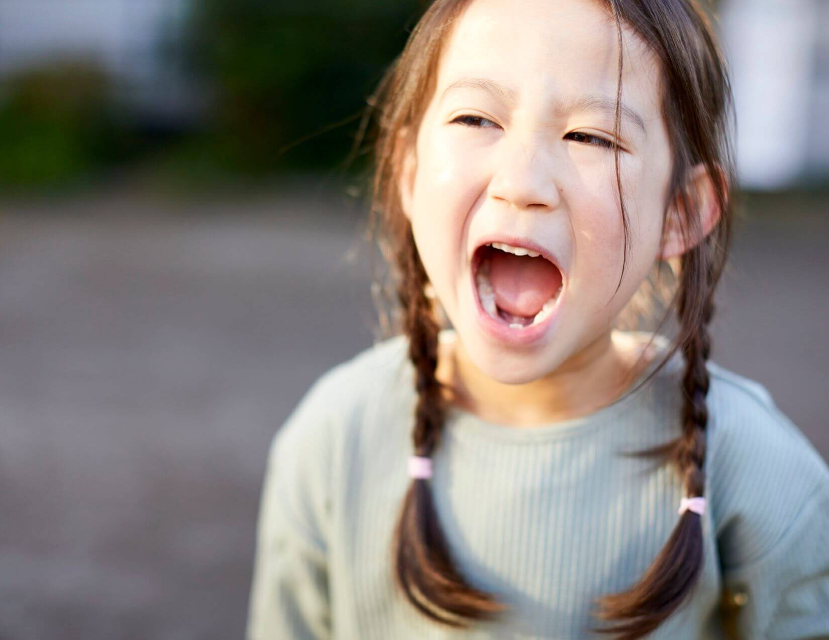 Young girl with light skin tone and brown braided pigtails with her mouth open wide.