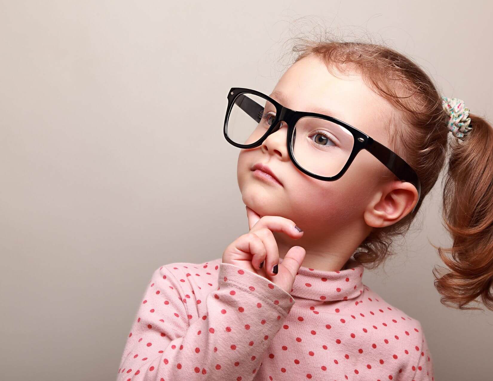 Young girl with light skin tone wearing large black glasses and looking to the side with her finger on her chin.