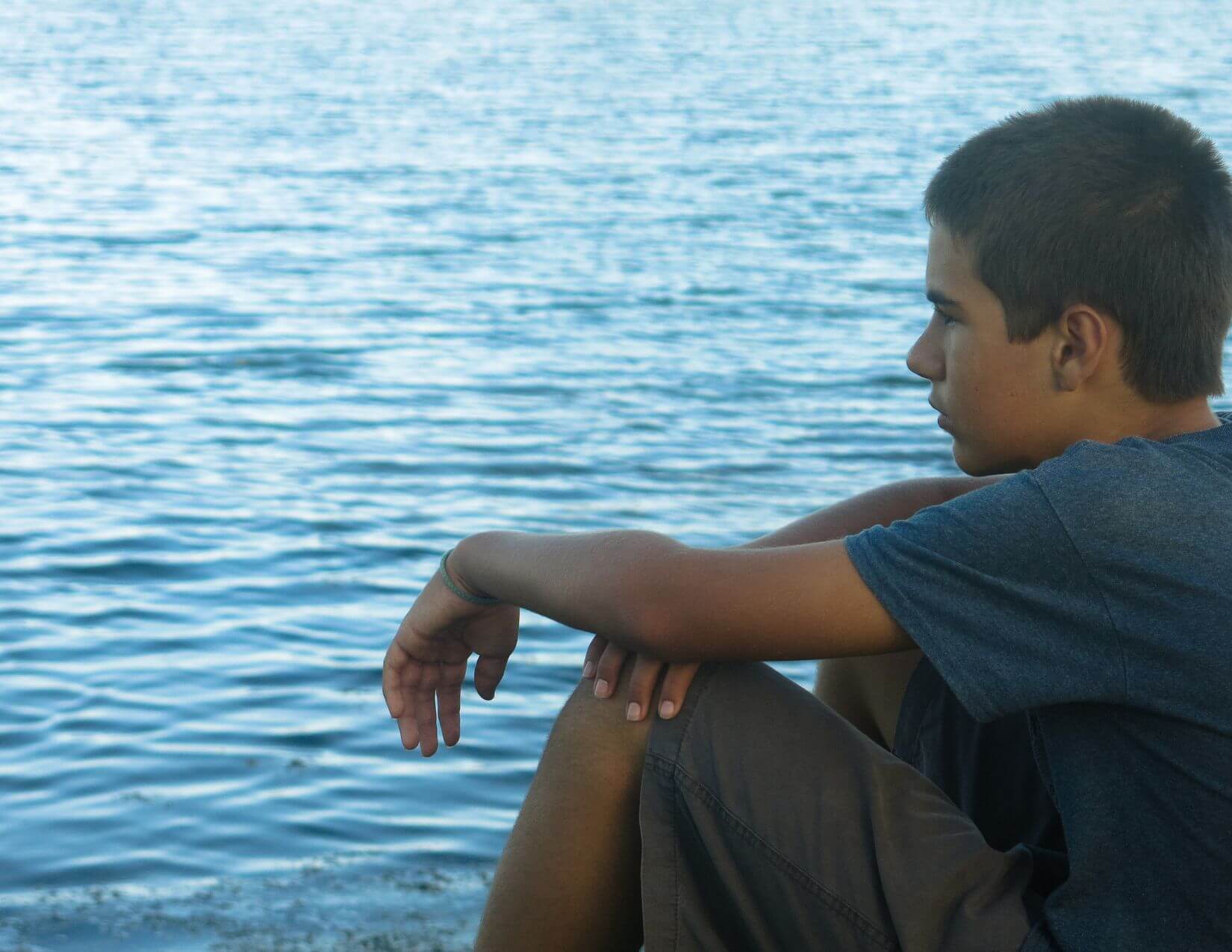 A middle school-aged boy sits looking out at calm lake water.
