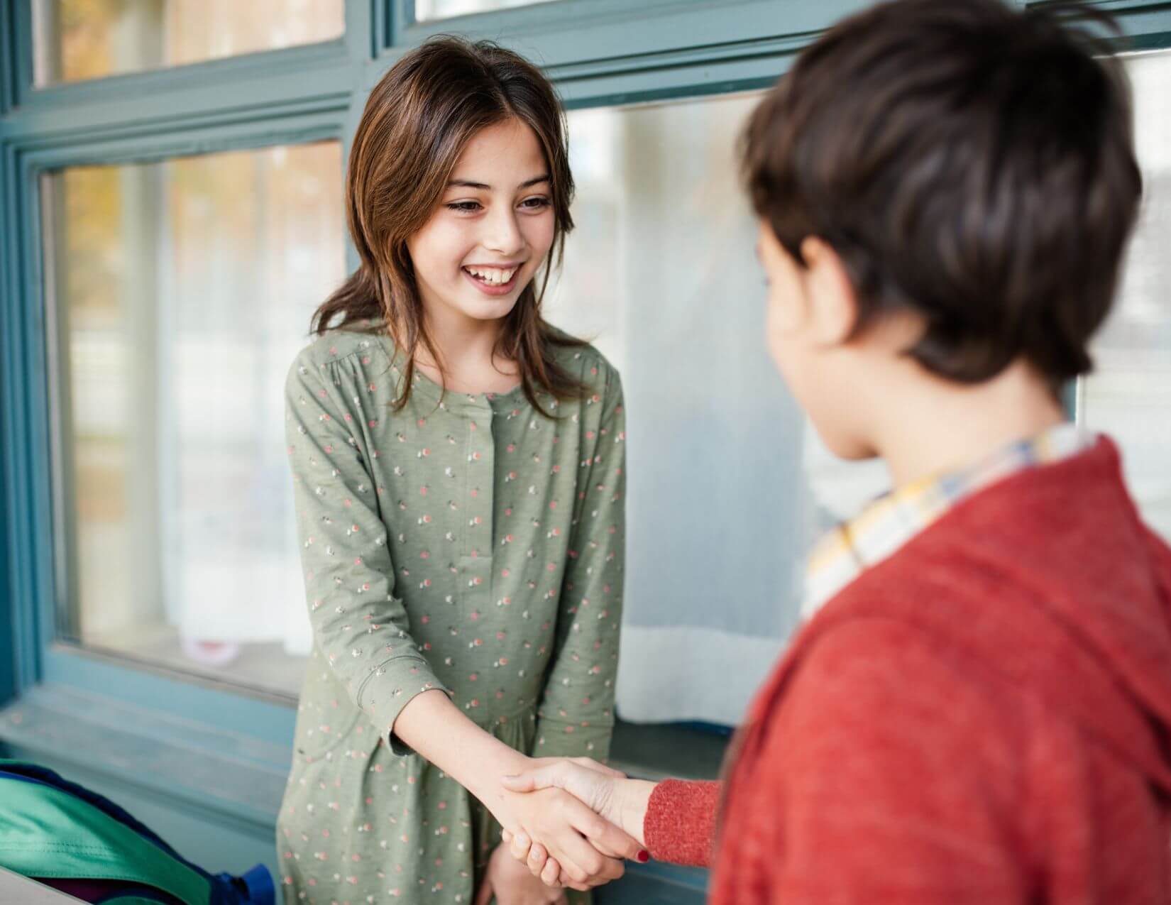 Two elementary-aged children with brown hair are shaking hands. The child facing the camera is smiling.