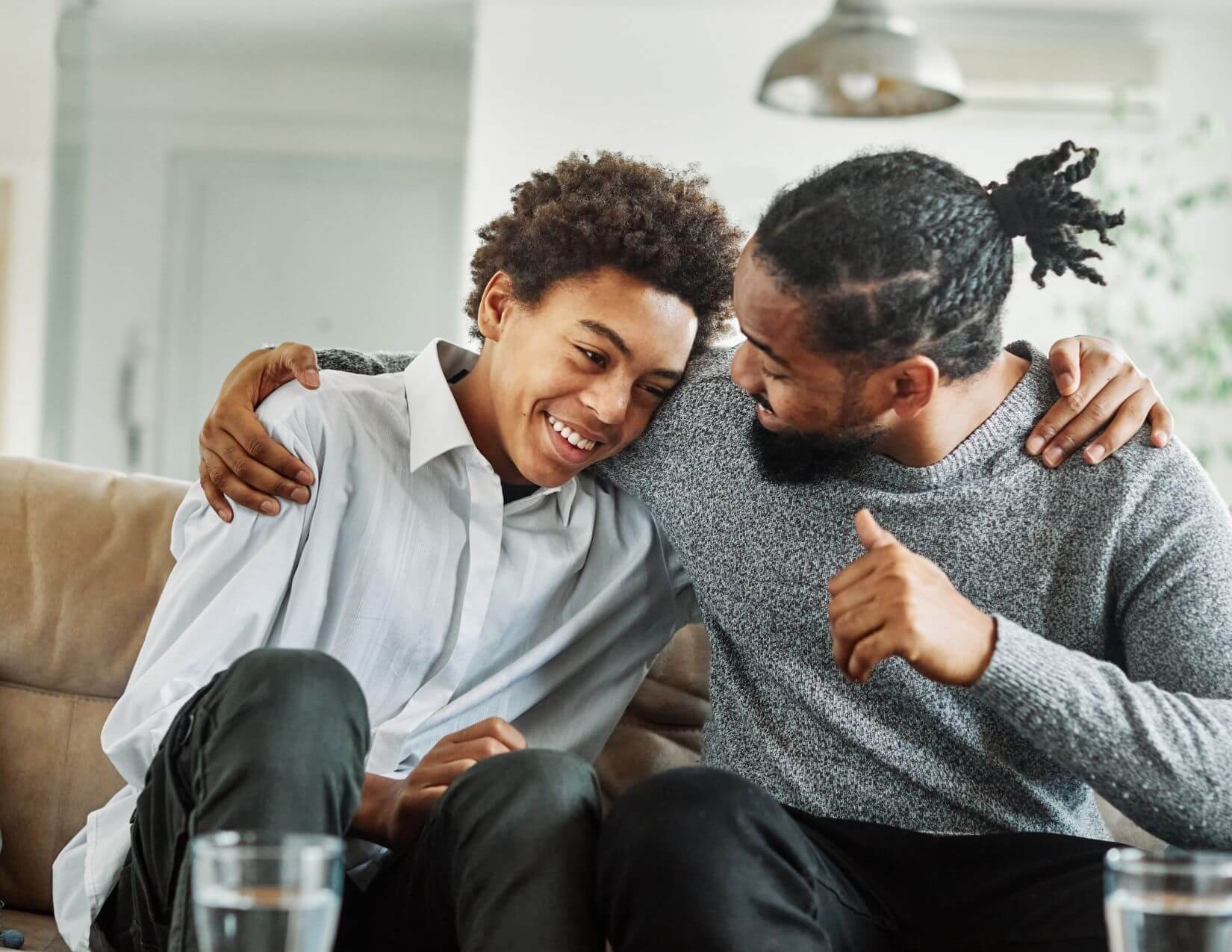 A man and young teen with darker skin tones sit on a couch in a sideways hug.