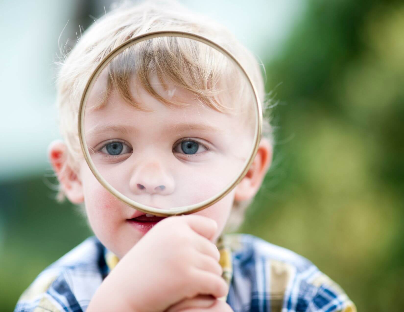 Toddler boy with blond hair looking through a big magnifying glass.