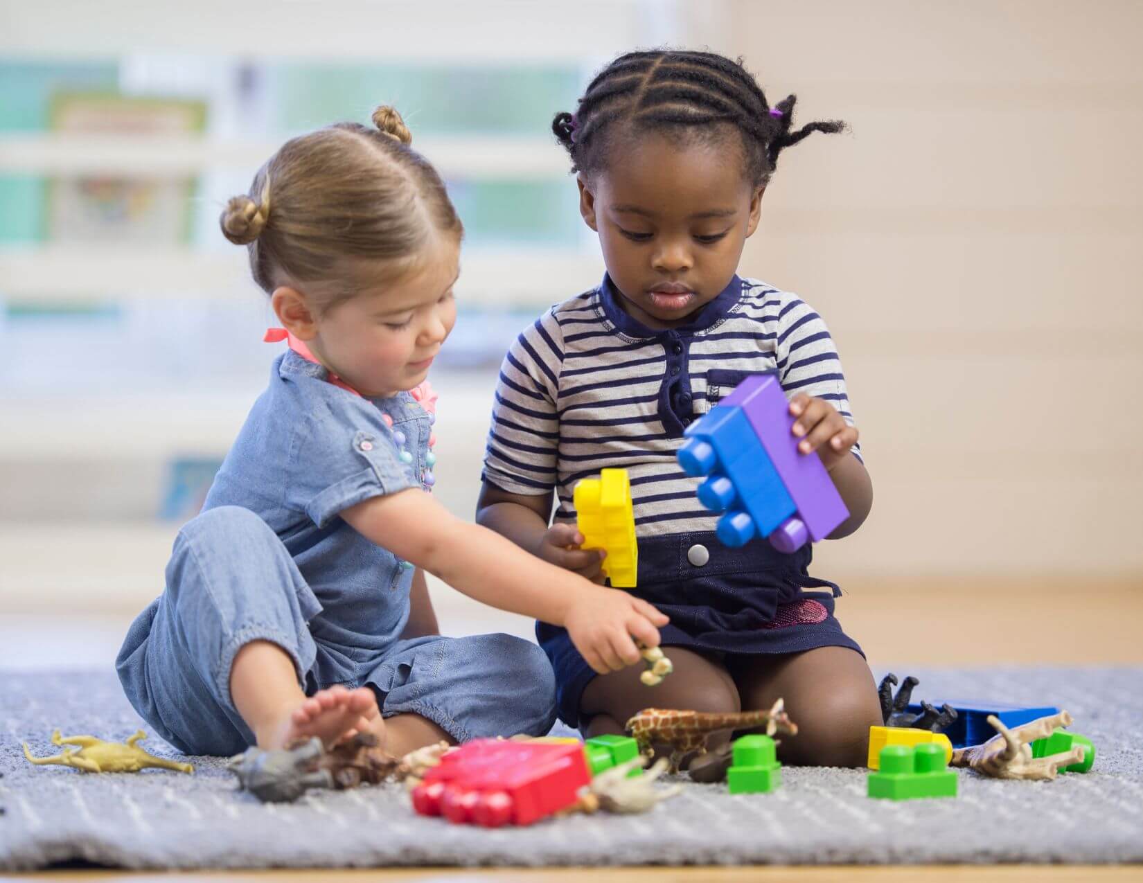Two little girls sit on a rug with toy animals and blocks. They are both looking at a toy giraffe on the rug.