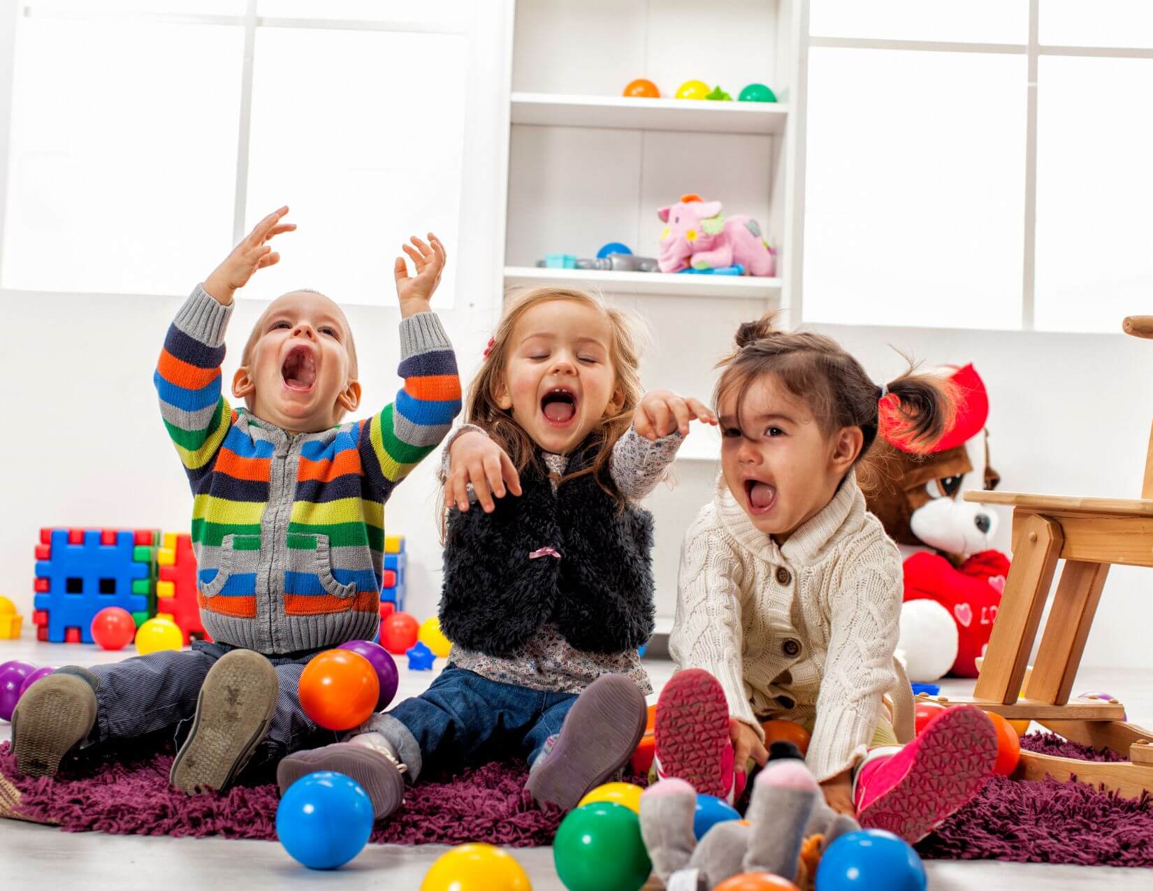 Three happy toddlers sit on a rug in a playroom with toys around them.